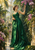 A woman in a long emerald-green dress reaches toward pink and lilac orchids in a lush glasshouse filled with palms.