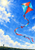 Red-and-cyan diamond kite with a long curling ribbon tail above sunlit clouds, flown by a small figure on a grassy ridge.