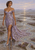A woman in a lavender floral gown stands in reflective tidal flats among purple flowers beneath a sunset sky.