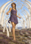 Woman in a purple dress stands beneath repeating pale bone arches in a rocky landscape.