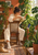 A woman in cream clothing waters abundant houseplants beside a bright window as a cat rests on the rug.