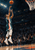 Airborne basketball player in a teal and white uniform releasing an orange ball toward a cropped hoop in a dark arena.