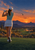 Woman golfer seen from behind with her club across her shoulders, overlooking a desert golf course at sunset.
