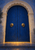 Closed cobalt-blue double doors with brass ring pulls beneath a scalloped pale stone arch, with warm light on the threshold.