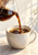 Dark coffee pouring from a tilted clear glass server into a large cream ceramic cup, with a ring of crema and bubbles.