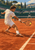 Tennis player in white slides low across a terracotta clay court to strike a yellow ball, with a net and distant opponent behind him.