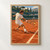 Tennis player in white slides low across a terracotta clay court to strike a yellow ball, with a net and distant opponent behind him.; frame shown for styling only