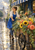 A smiling florist in a blue apron pushes a wooden cart filled with sunflowers and coral-pink flowers along a wet city sidewalk.