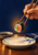 Chopsticks lift a salmon-and-cucumber maki roll above a patterned ceramic plate, with a soy dish and navy napkin.