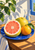 A cut pink-centered pomelo and two whole citrus fruits sit on a cobalt-blue plate beside a clear carafe under angular plant shadows.