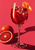 A ruby-red drink with ice and curled orange peel in a tall stemmed glass, beside whole and cut blood oranges on a pink-red ground.