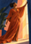Smiling woman in a flowing burnt-orange gown reaching to open an indigo curtain beside a bright window.