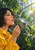 Profile of a woman in a yellow raincoat blowing an iridescent bubble through a brass wand in a leafy greenhouse.