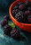 Glossy blackberries spill from a cropped persimmon-red bowl onto a dark teal tabletop beside a green leaf.
