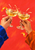 Two hands in blue and orange sleeves clink coupe glasses as golden liquid splashes between them against a red-orange background.
