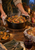 Steaming cast-iron casserole held with cloths over a wooden trivet amid harvest foods and tableware.
