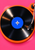 Glossy black vinyl record with a blue star-like label on an orange turntable against a pink background, with a cropped tonearm.