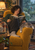 An adult with short dark curls reads in a mustard armchair beside a sleeping gray tabby and rain-streaked window.