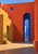 A cobalt-blue arched passage in a sunlit coral and ochre courtyard, with a circular window and diagonal shadow.
