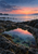 Reflective tidepool among dark coastal rocks at dawn, with coral-pink sky, calm sea, and sparse grasses.