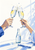 Two hands clink clear stemmed glasses with citrus slices above a table holding a clear bottle and folded napkin.