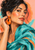Close portrait of a woman adjusting a glossy orange drop earring, wearing a turquoise scarf against a warm blush background.