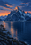 Snow-streaked mountain above dark reflective water, with a warm sunset reflection, distant peaks, rocks and small pines.