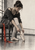 Clothed dancer seated on a studio bench and adjusting a ballet slipper, with a vivid red ribbon against a pale studio interior.