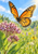 Orange-and-black monarch-like butterfly hovering above pink-purple flowers and green leaves against a softly blurred meadow and blue sky.