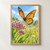 Orange-and-black monarch-like butterfly hovering above pink-purple flowers and green leaves against a softly blurred meadow and blue sky.; frame shown for styling only