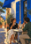 Two adults converse over drinks at a small café table beneath a cobalt awning and blue shutters on a sunlit street.