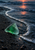 Translucent green triangular shard on wet black sand, encircled by a curved foamy wave with a coral reflection on the water.