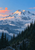 Snow-covered mountain with clouds around its slopes rises above dark evergreen forest beneath a coral sun.