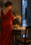 Adult woman in a red wrap dress lighting two candles at a dining table, with pears, a folded napkin, an empty chair, and a rainy doorway.