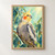 Yellow-faced cockatiel with an orange cheek patch perched on a branch among blue-green leaves against mint green.; frame shown for styling only