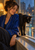 A woman in a blue robe sits beside a black cat on a windowsill before a rain-streaked window and blurred city lights.