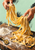 Two hands lift long golden pasta ribbons above a flour-dusted wooden board, with a brass pasta wheel at lower left.