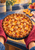 A golden lattice peach tart in a dark pan is held by red oven mitts as steam rises behind it.