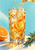 Amber-orange fizzy drink pours into a faceted glass with ice and orange peel, flanked by a cut orange and rosemary on pale aqua.