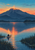 A glowing dawn summit above a lake, with a lone figure canoeing near shoreline reeds and coral-orange reflections on rippled water.