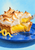 Lemon meringue tart with browned peaks on a cobalt-blue plate, a fork lifting a bite, and a lemon peel curl against pale cyan.