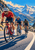 Three helmeted cyclists ride toward the viewer on a curving road beneath alpine peaks, with a lake behind them.