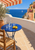 Cobalt-blue table with lemons, a carafe, and glasses on a sunny terrace overlooking the sea, with an olive tree, striped awning, and peach buildings.