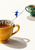 Tiny figure in a blue coat balances on a black line between an ochre teacup and a smaller green floral teacup on a warm-white background.