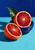 Two cut blood-orange halves in a cobalt-blue bowl, with a broad green leaf against a turquoise tabletop and indigo wall.