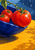Three red tomatoes, one attached to a leafy vine, sit in a deep cobalt bowl on a golden tabletop with rain droplets above.