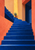 Broad cobalt-blue staircase rises through coral and apricot walls, turning at an amber-lit landing with a small potted agave.