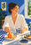 A laughing woman reaches for an orange while holding a patterned cup at a sunlit breakfast table with toast, blueberries, and leafy plants.