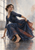 A fully clothed woman crouches in a flowing indigo-and-rust dress, gathering the skirt and turning over her shoulder beside a bright window.