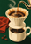 Cream pour-over coffee dripper and cup with flowing coffee, curling steam, rust-red cloth, and three coffee beans against dark green.
